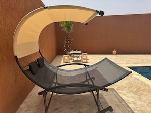 A reclining chair with a canopy, a small table, and chairs, near a pool in an outdoor setting.