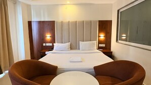Quarto de hotel com cama, duas poltronas marrons, uma mesa redonda pequena, um ventilador de teto e uma janela grande.