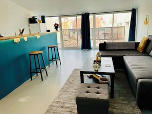 A modern living room with a sofa, ottoman, and a small table. There is a kitchen area with stools and a counter. Large windows with curtains are visible. - Panier Duplex — Terrace with Panoramic View of the Sea and the Rooftops of Marseille (Marseille)
