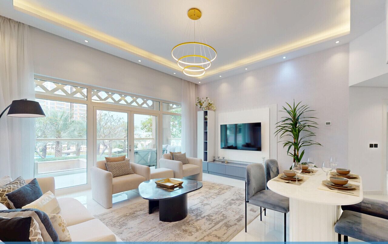 A modern living room with a flat-screen TV, a dining area with a round table, and large windows offering a view of the outdoors.