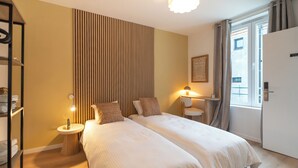 A hotel room with two beds, a bedside table with a lamp, a wooden floor, a window with curtains, and a door.