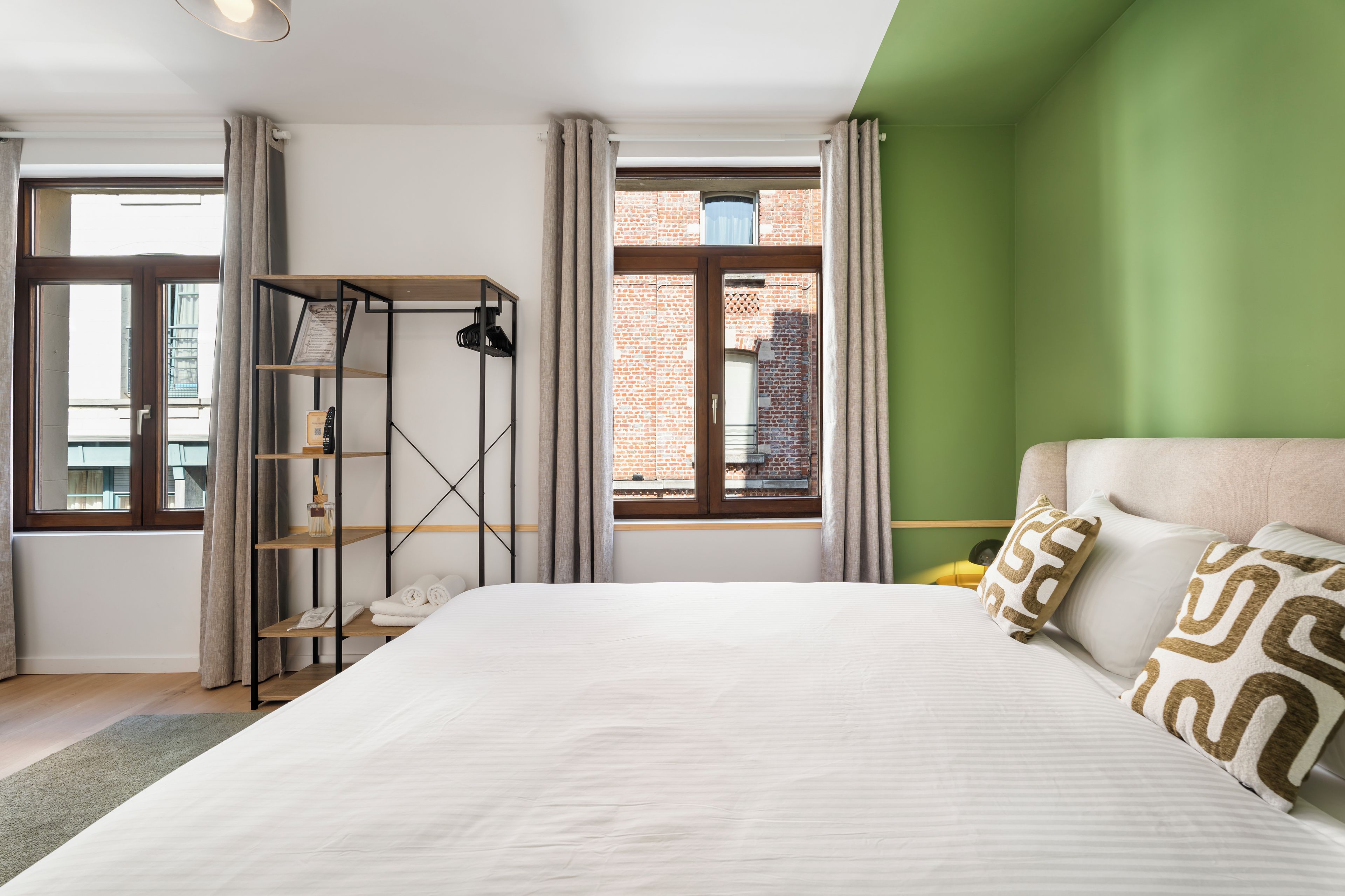 Ett hotellrum med en stor säng, en hylla i trä, utsikt över en byggnad med tegelstensfasad och en grön vägg.