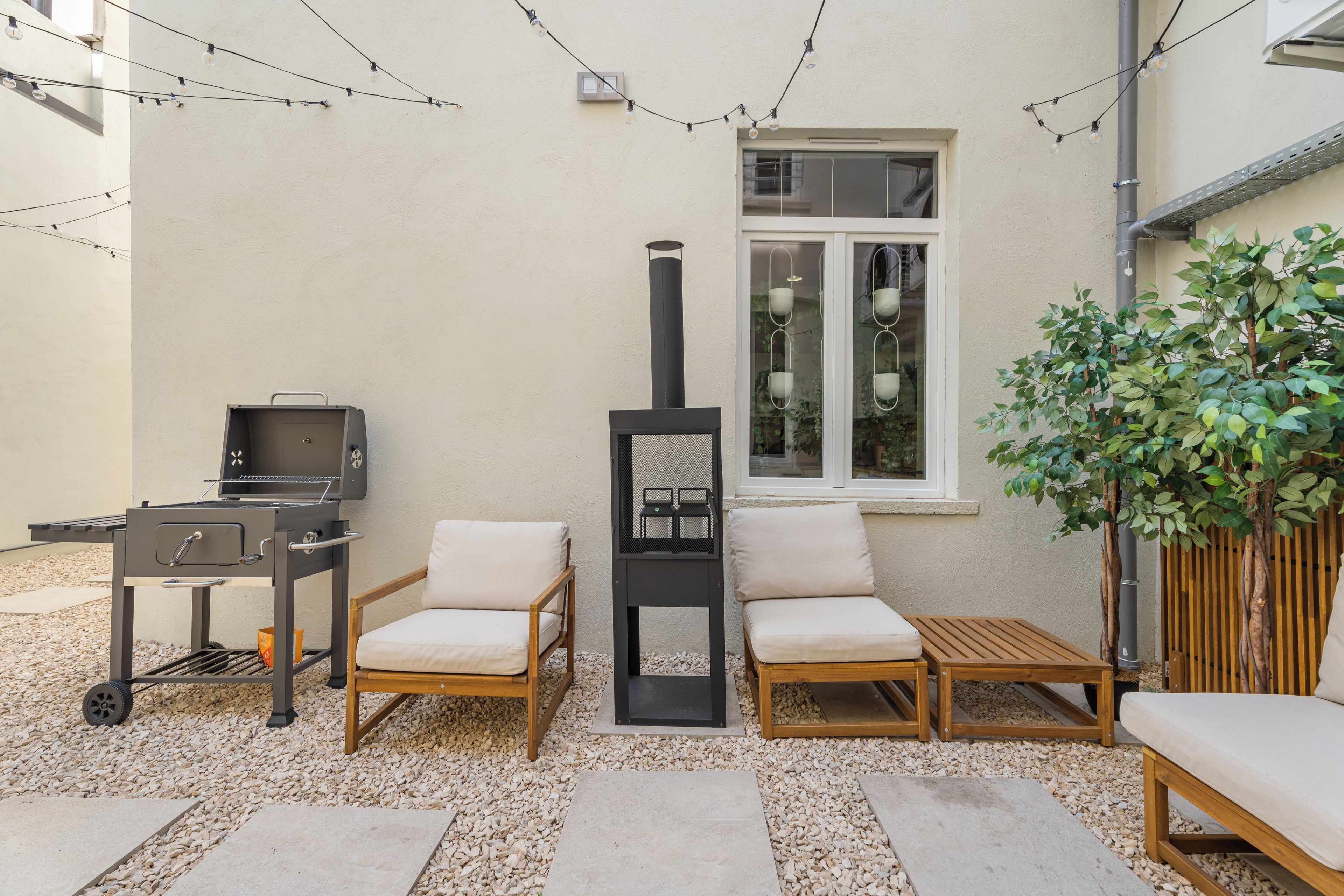 Une terrasse extérieure équipée d’un barbecue, de deux fauteuils confortables, d’une petite table, d’une plante en pot et d’une fenêtre donnant sur un espace intérieur aménagé avec des plantes.