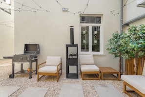 Un patio exterior con una parrilla, dos sillas acolchadas, una mesita, una planta en maceta y una ventana con vistas a plantas de interior.
