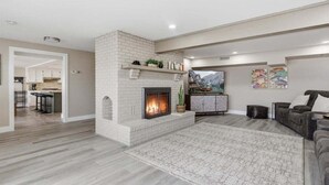 A living room with a fireplace, a sofa, a rug, and a kitchen in the background. - Outdoor Dream Lakefront Home w/ Private Dock, 2 Slips & Kayak (Sunrise Beach)