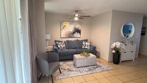 A living room with a grey sofa, a small coffee table, a ceiling fan, a painting on the wall, a mirror, and a cabinet with a vase of flowers. - Recently Renovated Marvelous 3-bedroom condo with AC in charming Aquatika, Loiza (Loiza)