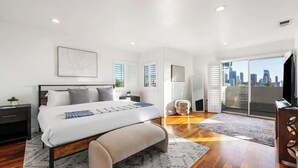Ein modernes Schlafzimmer mit Bett, Nachttischen, einer Bank, Stadtblick durch große Fenster und einem Balkon.