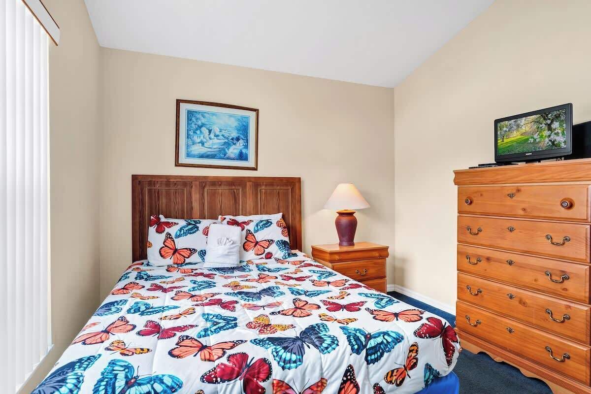 A bedroom with a bed, a wooden dresser, a nightstand, a lamp, a television, and a framed picture on the wall.