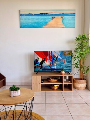 A living room with a television displaying a sailing race, a wooden table, a potted plant, and a painting of a pier. - 2-Bedroom. Stunning panoramic ocean view - Terraces & BBQ, Sainte-Luce (Sainte-Luce)