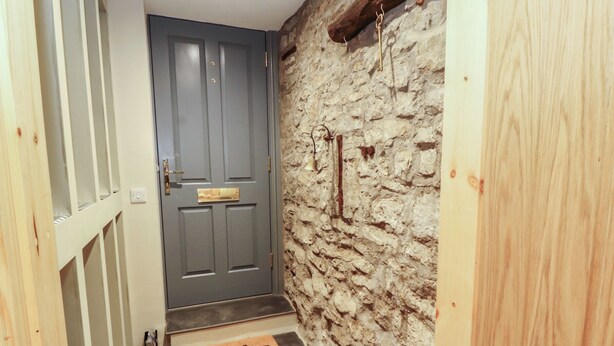 Een smalle gang met een stenen muur, een houten deur met een messing knop, en een klein houten bankje met een kleed.