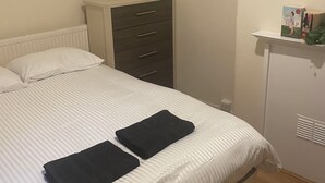 Una cama con una colcha a rayas blancas, dos toallas negras, un aparador gris y un estante pequeño con libros.