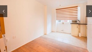 Una habitación con suelo de madera, paredes blancas, una ventana con persianas y una zona de cocina con microondas, además de una puerta.
