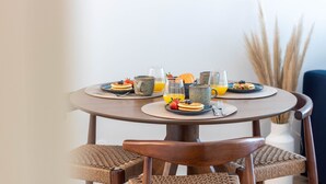 Una mesa redonda con platos de tortitas y fruta, dos vasos de zumo de naranja, y un jarrón con hierbas altas al fondo.
