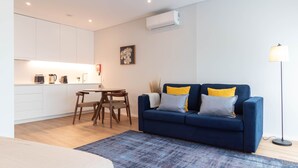 Una sala moderna con un sofá azul, una pequeña mesa de comedor y una zona de cocina con armarios blancos.
