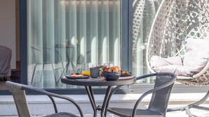 Una mesa redonda con alimentos para el desayuno, dos sillas, una silla de mimbre y una puerta de cristal con cortinas.