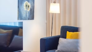 A modern living room with a flat-screen TV, a dark blue sofa with a yellow and gray pillow, and a floor lamp. - 1T | Rosário 244 by LovelyStay (Porto)