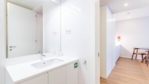 A modern bathroom with a white sink, mirror, and cabinets, a wooden floor, and a small table with chairs in the adjacent room. - 1T | Rosário 244 by LovelyStay (Porto)