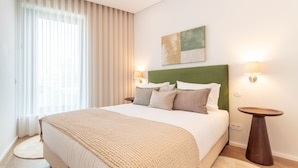 A hotel room with a bed, a headboard, two bedside lamps, a painting on the wall, and a window with curtains. - 1T | Rosário 244 by LovelyStay (Porto)