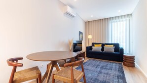 A modern living room with a round wooden table, two wicker chairs, a dark blue sofa with yellow cushions, a flat-screen TV, and a large window with sheer curtains. - 1T | Rosário 244 by LovelyStay (Porto)