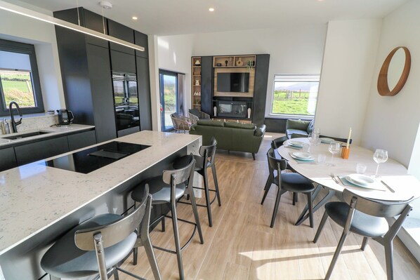 A modern kitchen with a large island, bar stools, a dining table with chairs, a sofa, and a TV mounted on the wall.
