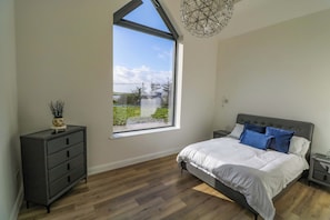 A bedroom with a large window, a bed with white bedding, a dark dresser, and a modern chandelier.