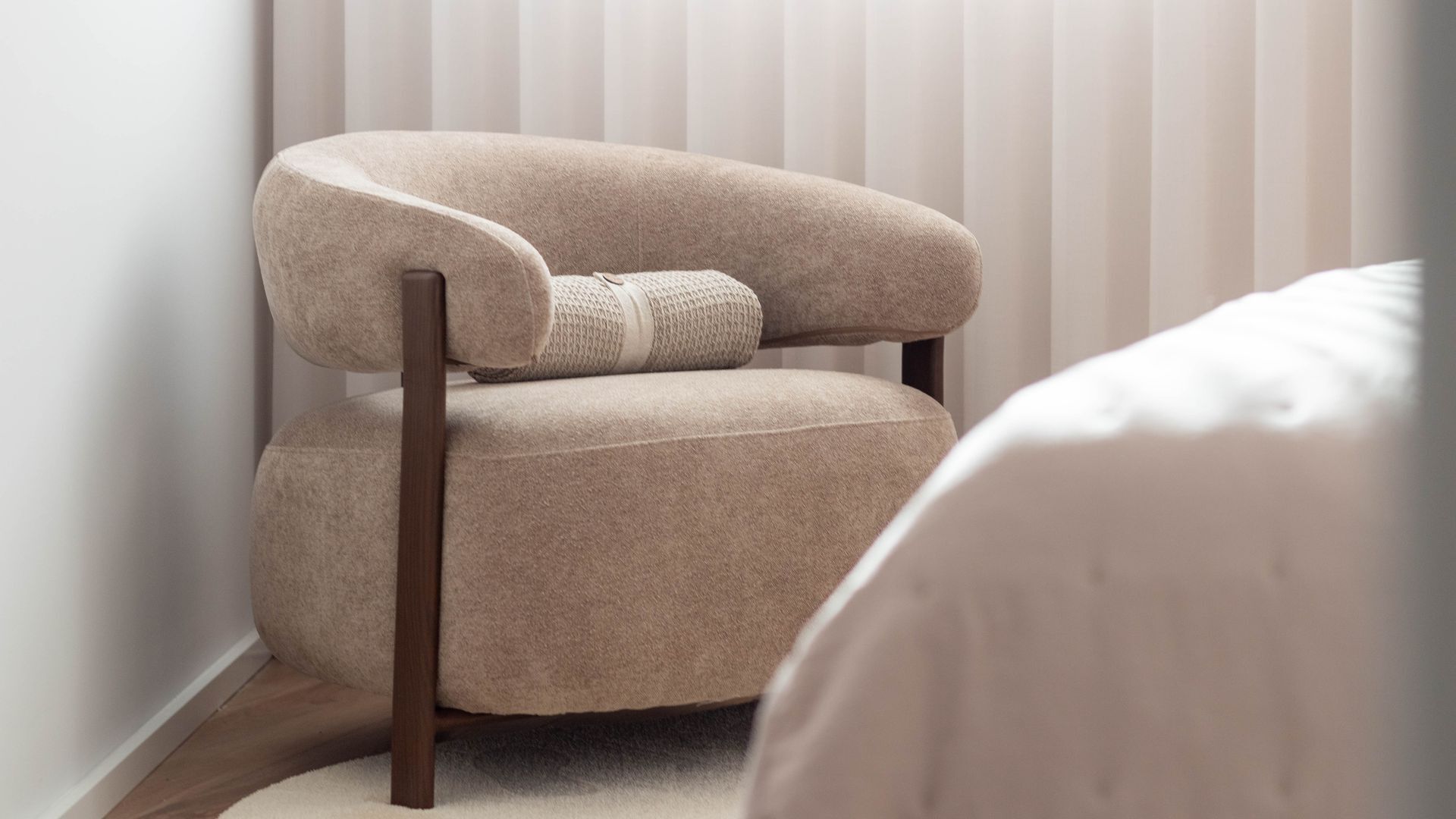 A corner view of a room with a beige armchair, a wooden floor, and a white wall with vertical blinds.