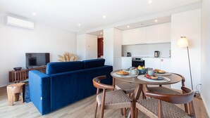 Un salon moderne avec un canapé bleu, une table à manger entourée de chaises en osier, un téléviseur à écran plat et un coin cuisine.