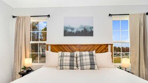 A bedroom with a large bed, two bedside lamps, a headboard with a herringbone pattern, and a window with curtains. - Cute Condo | Hot Tub | Minutes to Pigeon Forge! (Sevierville)