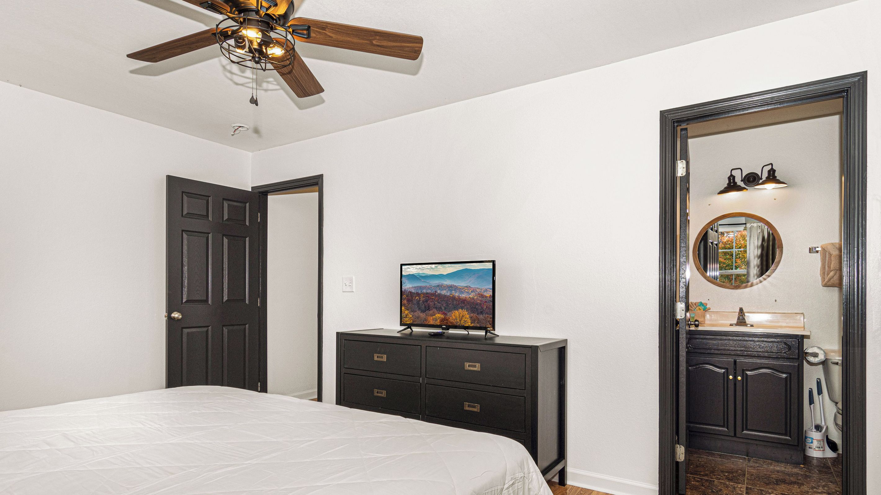 A bedroom with a bed, a dresser, a television, a ceiling fan, and a bathroom visible through an open door.