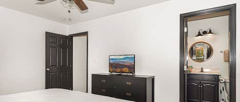 A bedroom with a bed, a dresser, a television, a ceiling fan, and a bathroom visible through an open door.