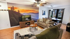 A living room with a large TV, a sofa, a coffee table, and a ceiling fan. - Cute Condo | Hot Tub | Minutes to Pigeon Forge! (Sevierville)