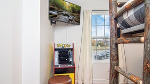 Une pièce équipée d’une borne d’arcade Pac-Man, d’un téléviseur mural diffusant des images de la nature et d’une fenêtre avec des rideaux.