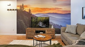 A living room with a flat-screen TV on a wooden stand, a sofa, a coffee table with magazines, and a large wall mural of a forest and mountains. - Cute Condo | Hot Tub | Minutes to Pigeon Forge! (Sevierville)