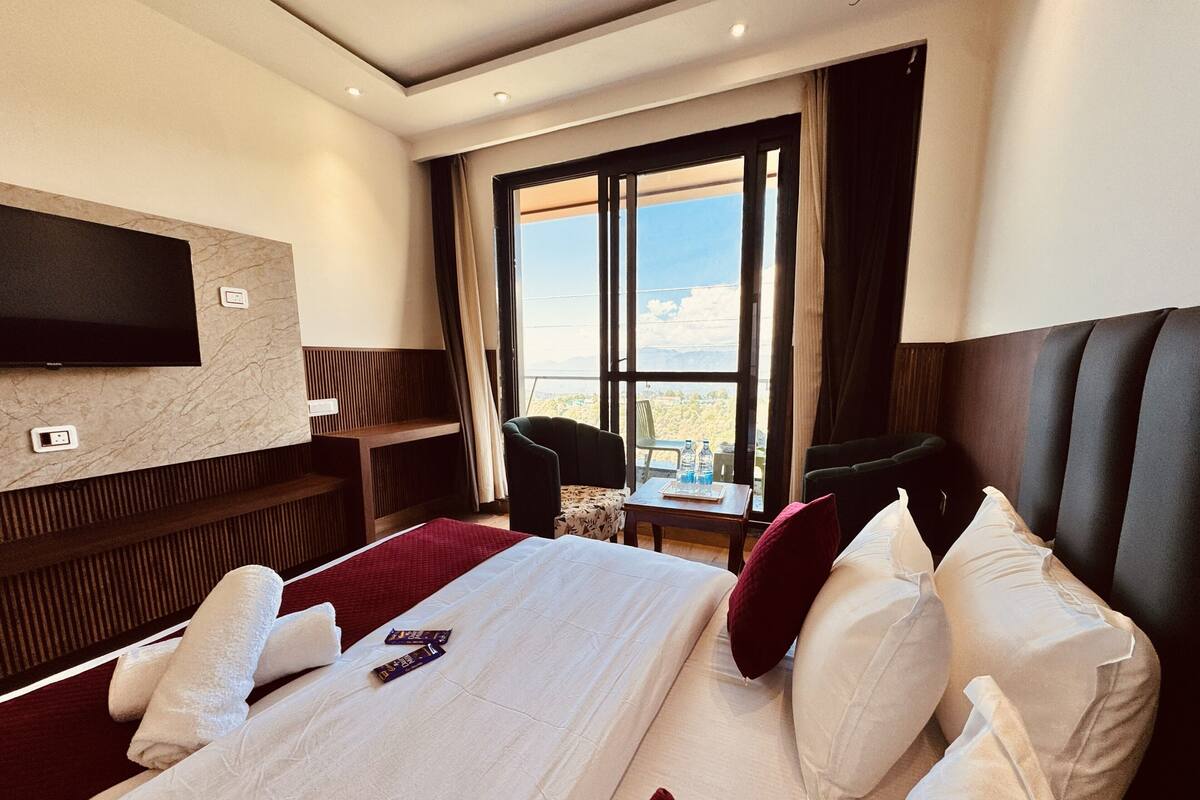 A hotel room with a large bed, a TV mounted on the wall, a small table with a chair, and a window with a view.
