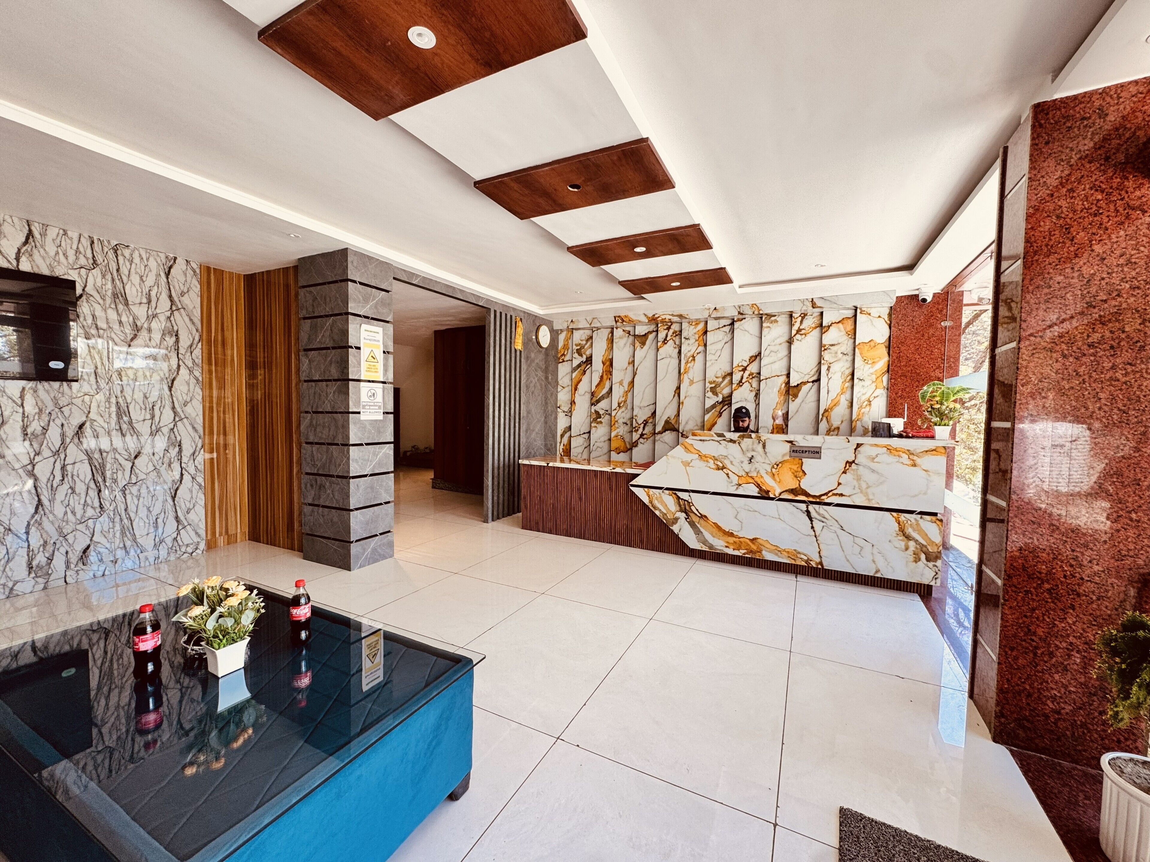 Un hall d’hôtel avec un comptoir de réception en marbre, une table basse en verre bleu ornée de fleurs et d’une bouteille, et un design moderne au plafond.