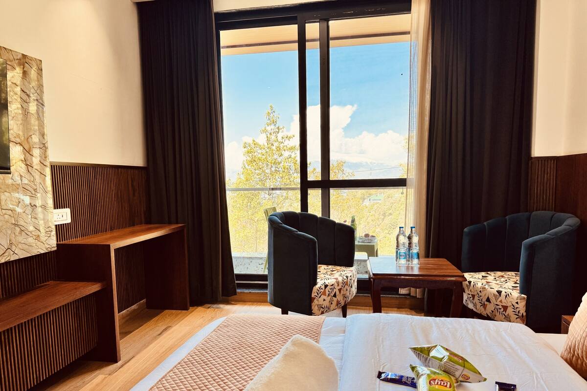A hotel room with a bed, two armchairs, a small table, a bench, and a large window with a view.