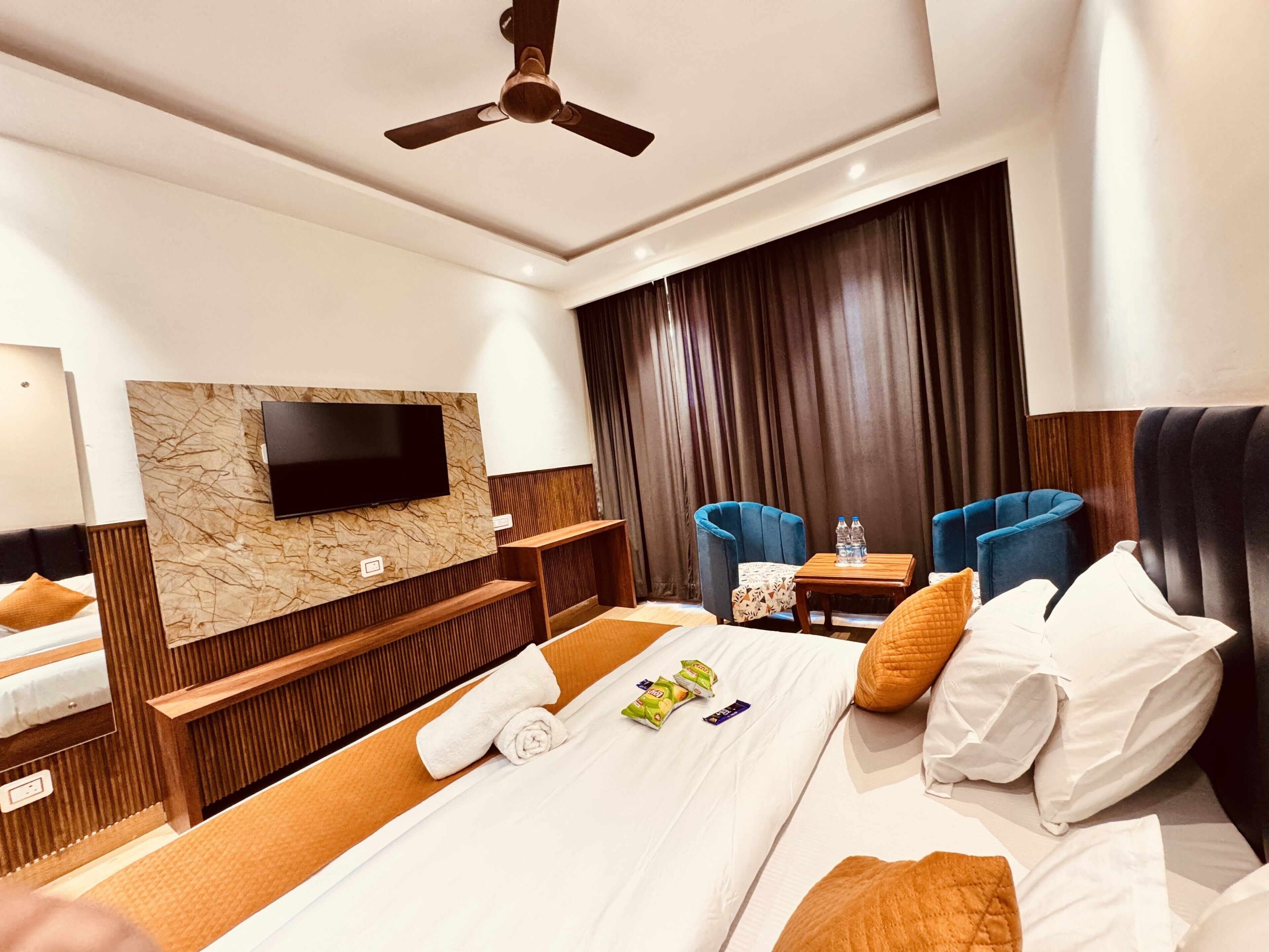 Une chambre d’hôtel avec un lit, un téléviseur fixé au mur en marbre, un ventilateur de plafond, une petite table avec des bouteilles d’eau et deux fauteuils bleus.