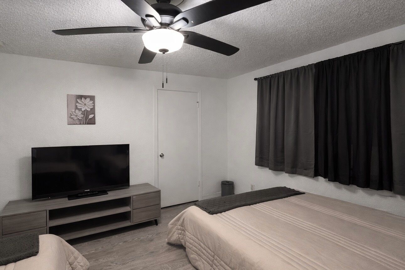 A bedroom with a bed, a TV on a stand, a ceiling fan, a door, and a window with curtains.