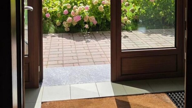 A door leading to a room with a view of a mountain, a garden with pink flowers, and a house in the background. - Albergo Ristorante Casa Corti (Valbondione)