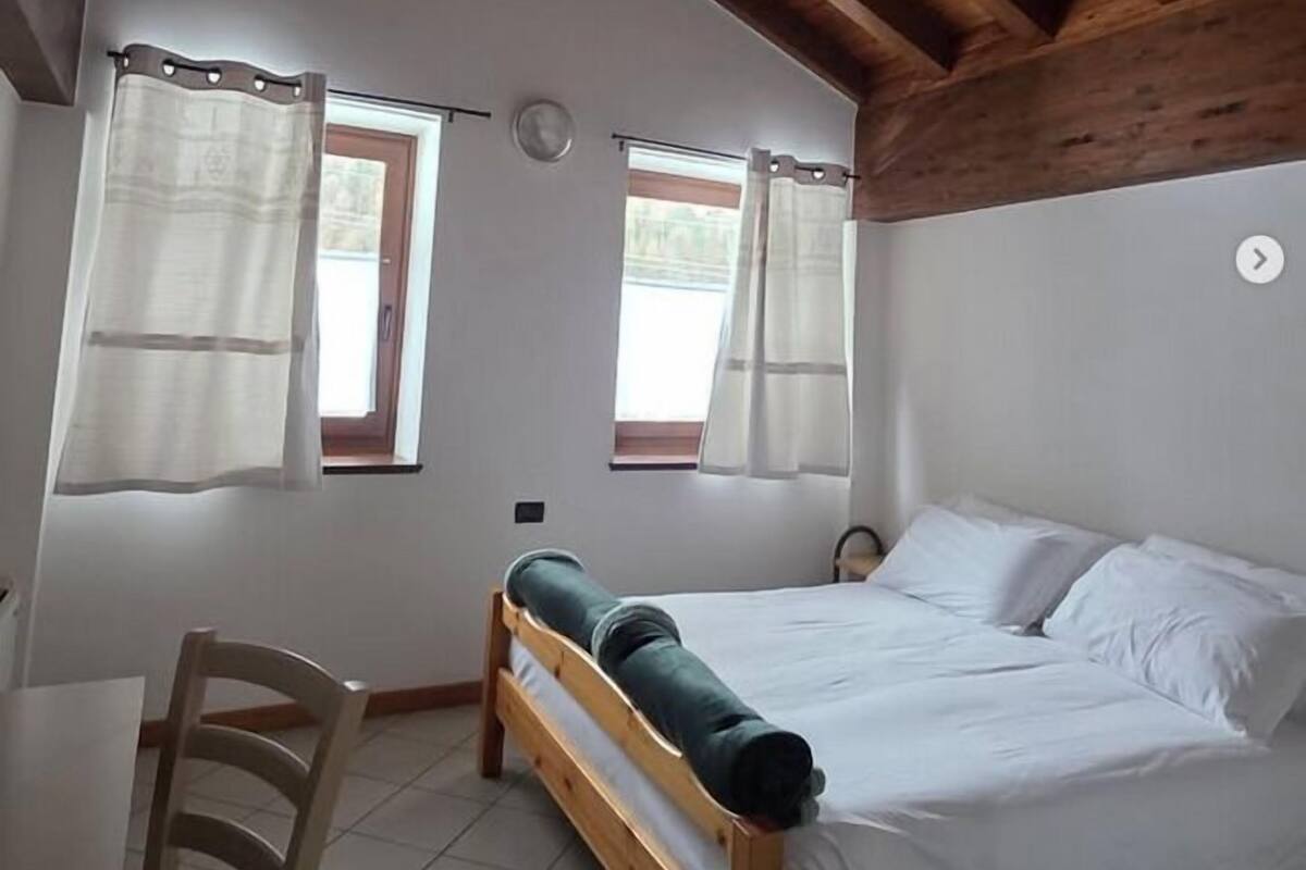 A bedroom with a wooden ceiling, two windows with curtains, a bed, a chair, and a small table.