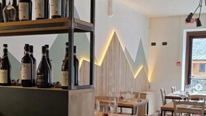 A restaurant interior with wooden tables and chairs, a wine rack with bottles, and a modern light fixture. - Albergo Ristorante Casa Corti (Valbondione)