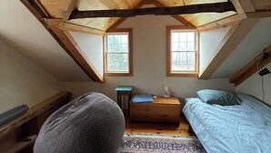 Een kleine slaapkamer op zolder, met schuine wanden, twee ramen, een bed, een bureau en een relaxfauteuil.
