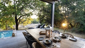 A wooden deck with a dining table set for two, surrounded by wicker chairs, a lantern, and a pool in the background. - Villa Riviersig,  a new stylish bushvilla (Hoedspruit)