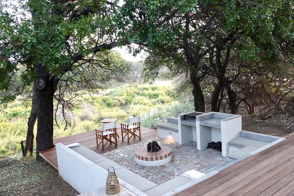 An outdoor seating area with a stone fire pit, wooden deck, and a view of a natural landscape. - Villa Riviersig,  a new stylish bushvilla (Hoedspruit)