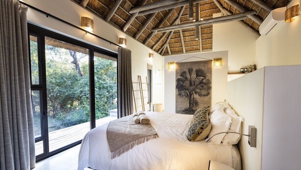 A bedroom with a large bed, a view through a sliding door, a thatched ceiling, and a wall-mounted artwork. - Villa Riviersig,  a new stylish bushvilla (Hoedspruit)
