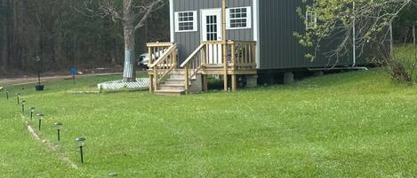 A small, single-story building with a porch and steps, surrounded by a grassy area and trees.