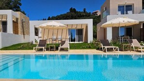 A modern pool area with lounge chairs and umbrellas, surrounded by a stone building and a glass structure. - Arillas Peaceful Bungalows (Corfu)