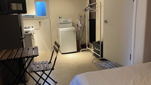 A compact hotel room with a bed, a small table, a folding chair, a microwave, a refrigerator, a washing machine, and a shower area.