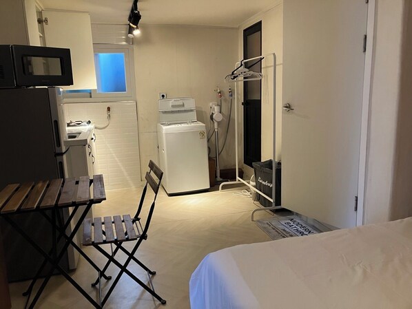Camera d'albergo compatta con un letto, un piccolo tavolo, una sedia pieghevole, un forno a microonde, un frigorifero, una lavatrice e un angolo doccia.