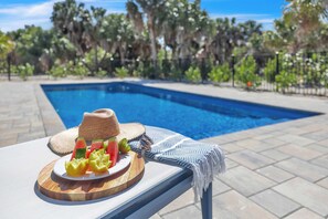A poolside setting with a table featuring a plate of fruit, a straw hat, and sunglasses. - New Build | Pool · Outdoor Bar · 2 Golf Carts Incl. (Captiva)